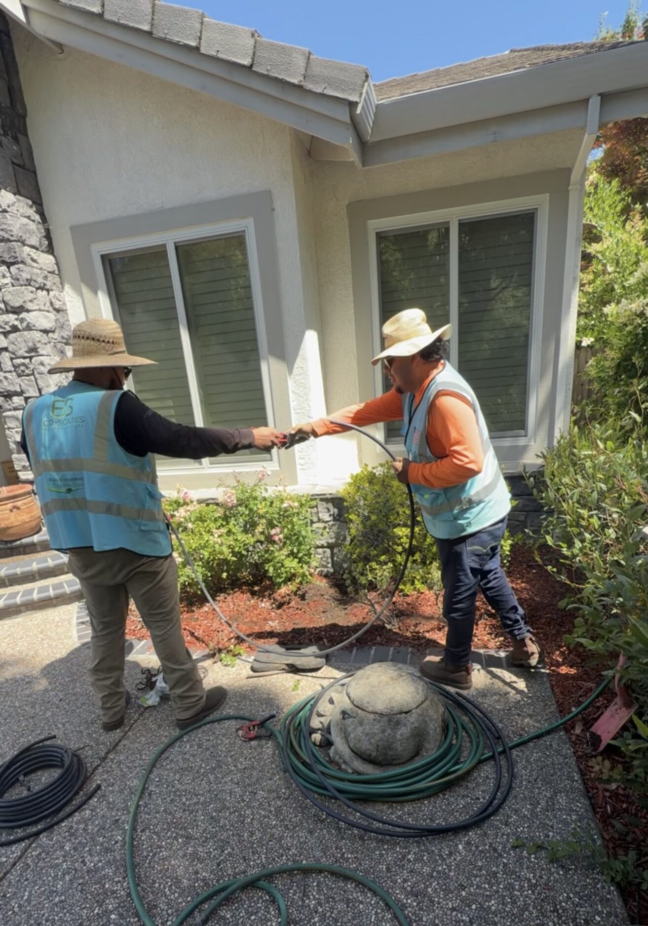 Two crew members passing irrigation tubing along a front-entry planting bed with hoses coiled on the walkway