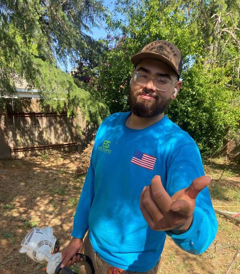 Eco Scapes owner Carlos in a blue Eco Scapes shirt and safety glasses holding a string trimmer in a backyard