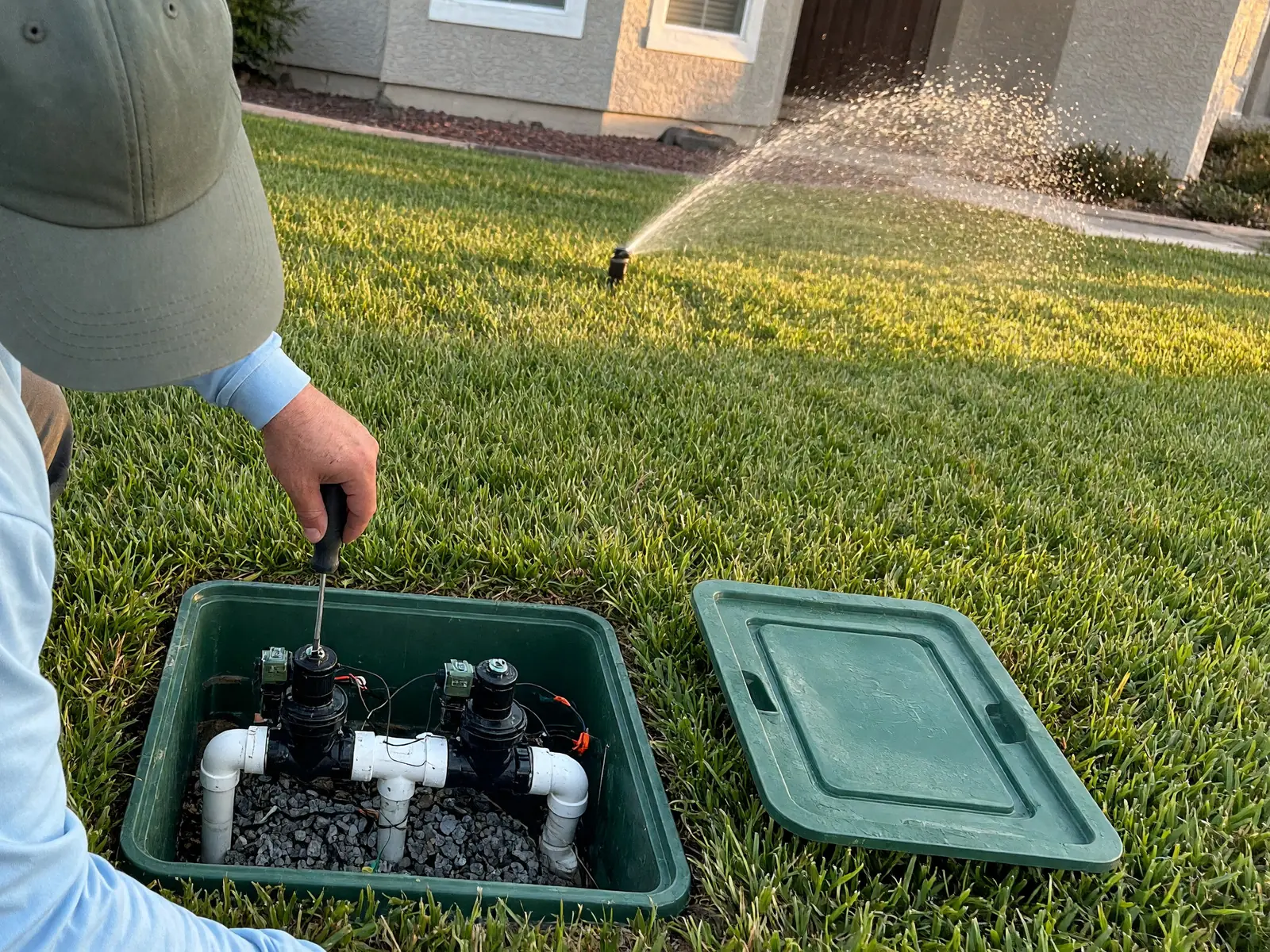 A technician in a light blue long-sleeve shirt and cap works on two sprinkler valves inside an open in-ground valve box on a front lawn while a rotor sprinkler waters the lawn behind.