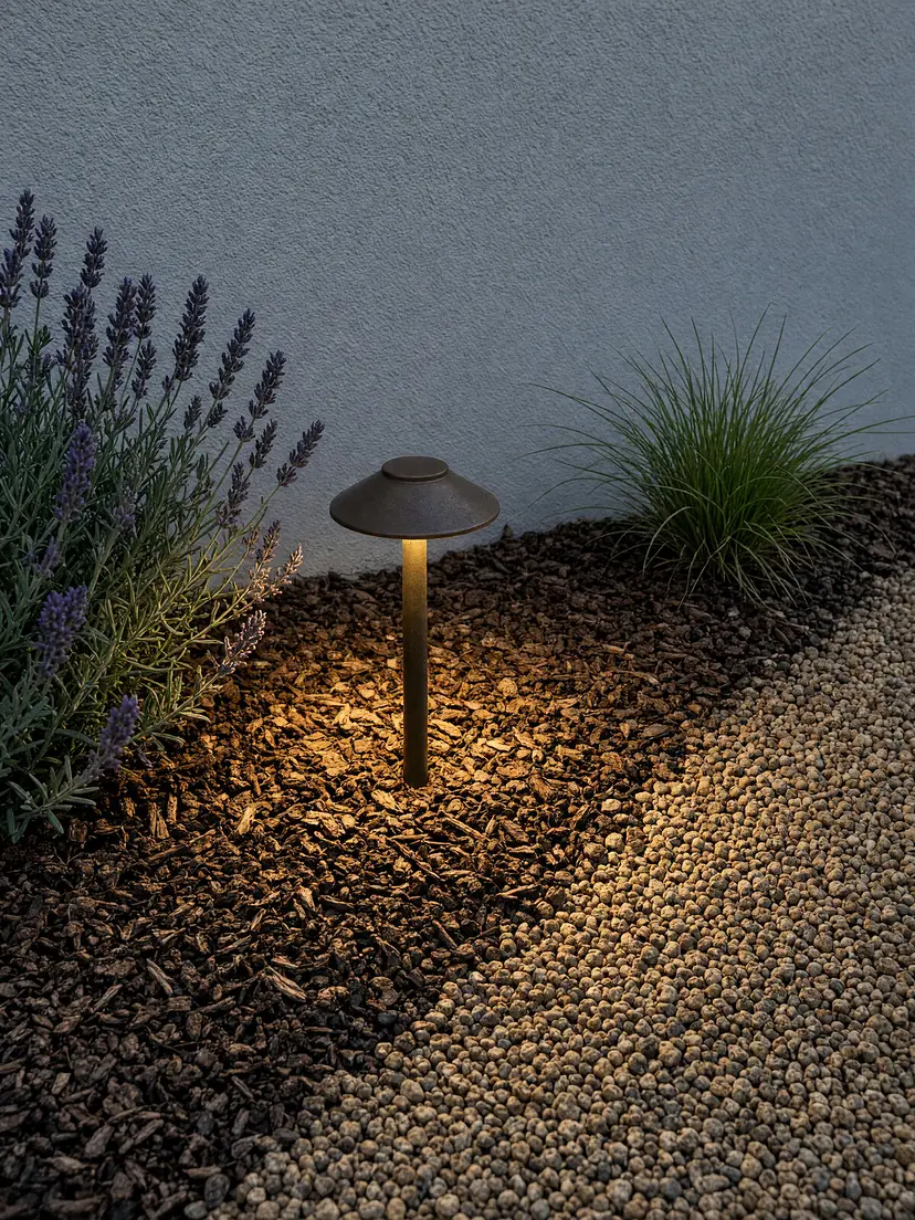 A bronze path light glowing over bark mulch and pea gravel beside lavender and an ornamental grass at dusk.