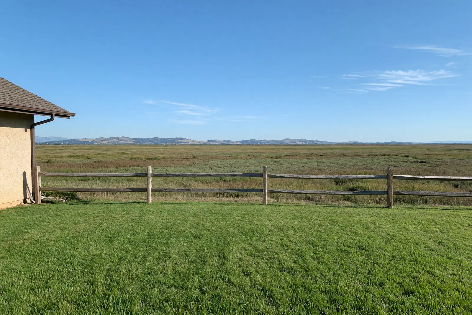 Illustration of a mowed back lawn ending at a split-rail wooden fence, with flat marsh grassland beyond and low hills under a wide afternoon sky.
