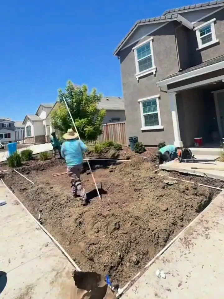 Crew member in a straw hat carrying a long-handled tool across the bare soil of a front yard being prepared for planting