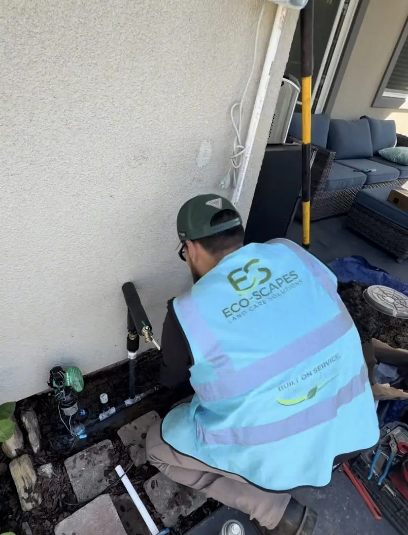 Technician in an Eco Scapes vest working on an insulated supply riser and hose bib against a house wall, with irrigation valves at its base