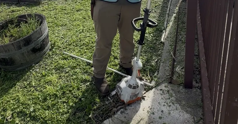 String trimmer resting on a freshly trimmed side-yard lawn along a metal fence, with a half-barrel planter nearby