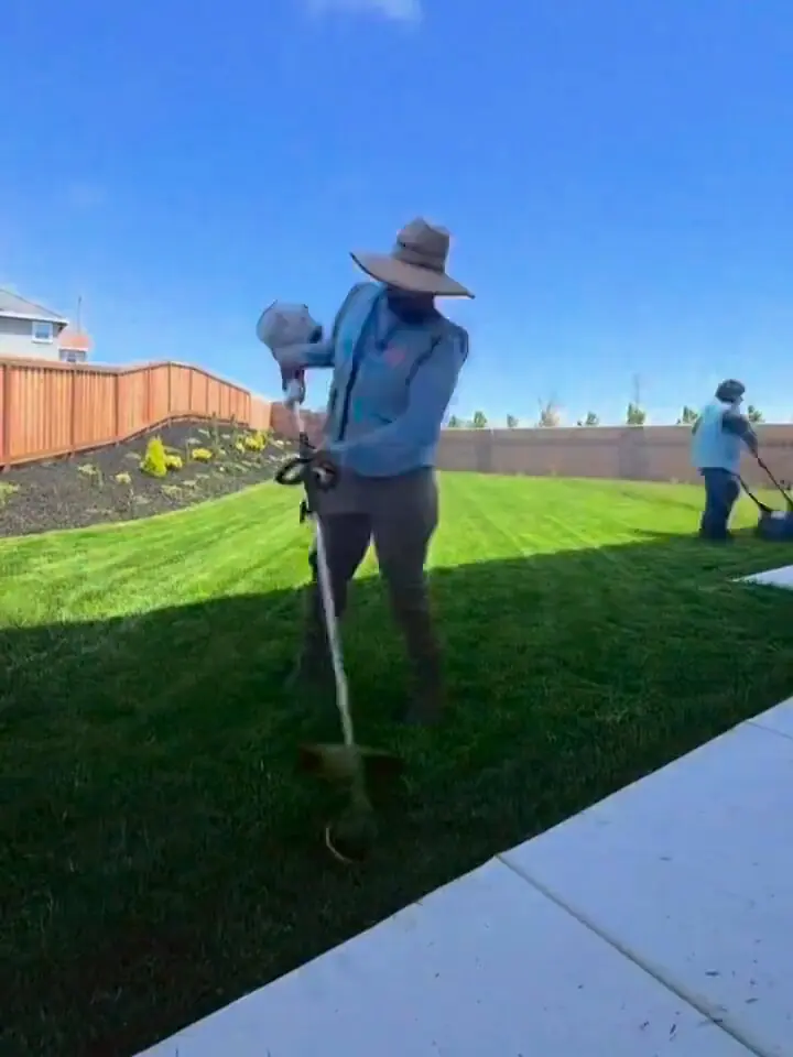 Crew member in a wide-brim hat running a string trimmer on a backyard lawn while a teammate mows behind him